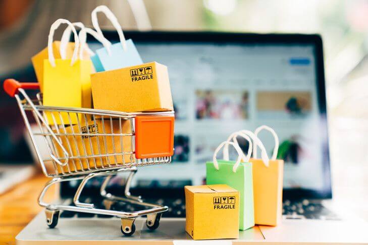 Image représentant un mini caddie rempli de colis et de sacs colorés devant un ordinateur portable, symbolisant les ventes en ligne et le business des influenceurs